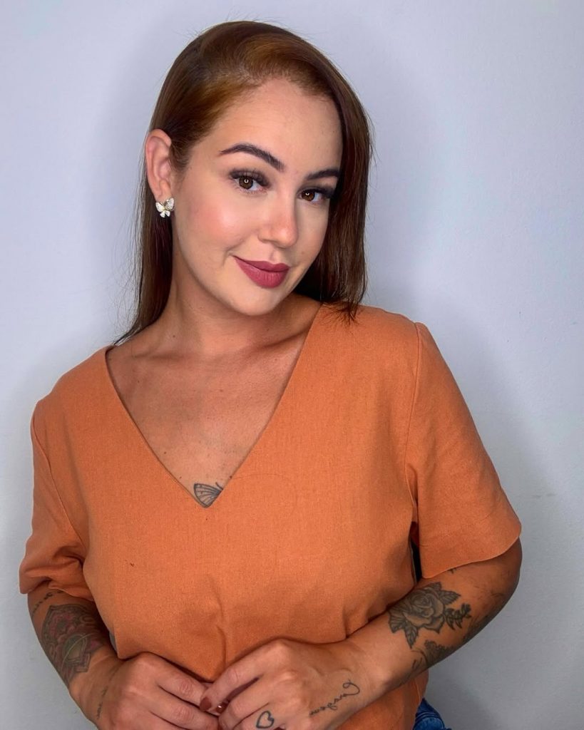 Eduarda Rafaella posa com um sorriso de lado, usando uma camiseta laranja e em um fundo branco. Ela possui tatuagens nos braços